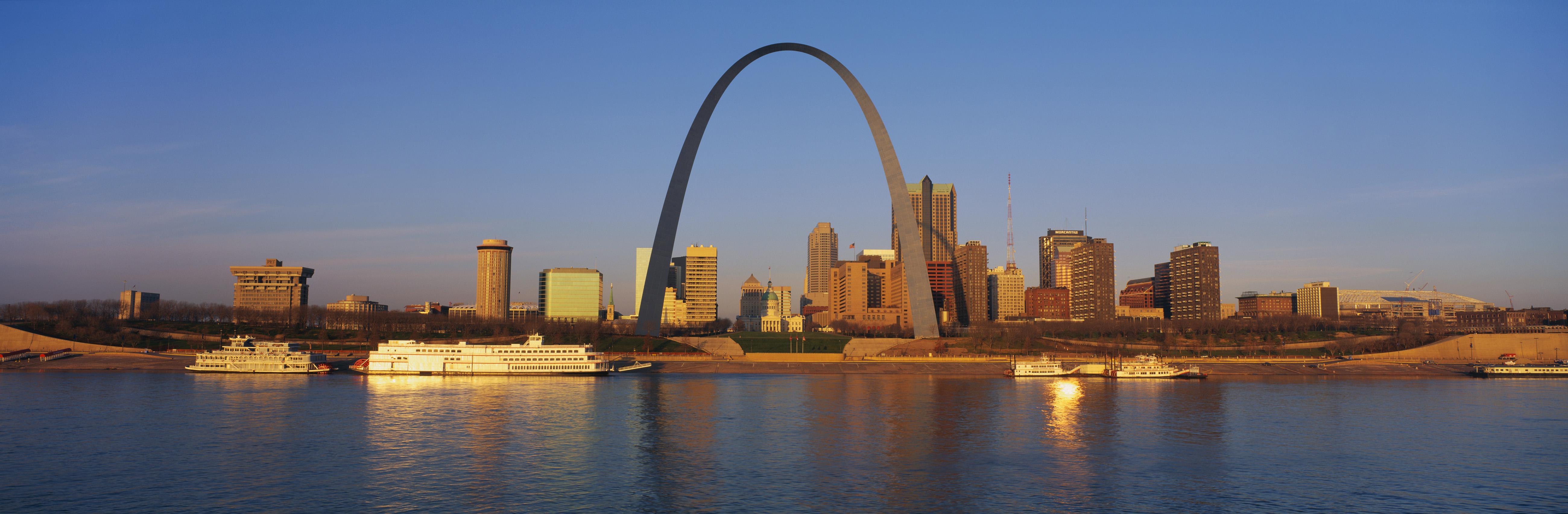 This is the skyline at sunrise. It is situated along the Mississippi River. There are riverboats on the water with the St. Louis Arch in clear view.